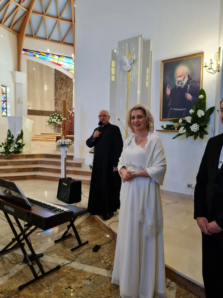 Renata Drozd w białej sukni i kapłan podczas uroczystości w kościele, obok instrumentu klawiszowego.